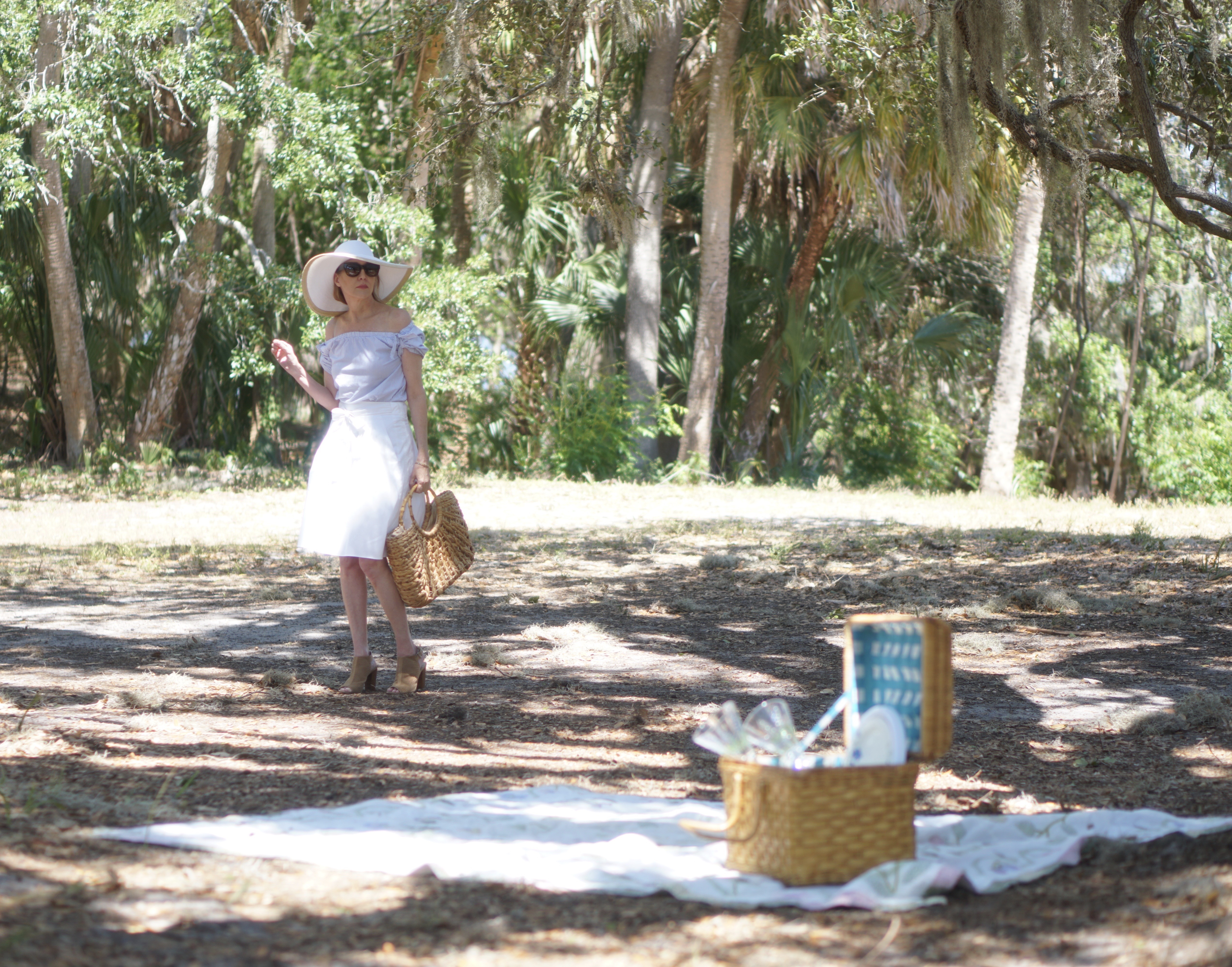 Woman standing near picnic basket in wide brimmed hat white skirt and blue top