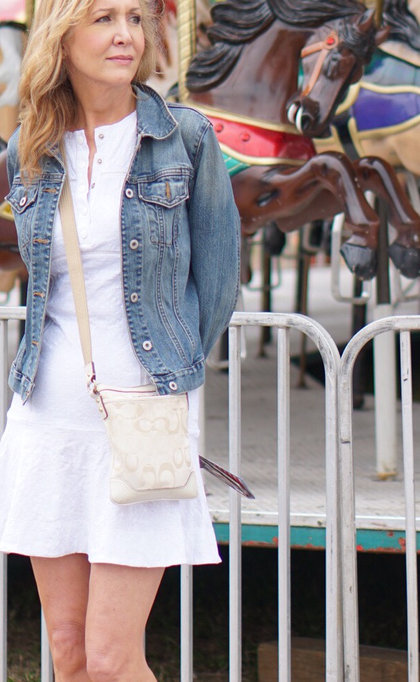 Woman standing next to carousel wearing white dress and denim jacket