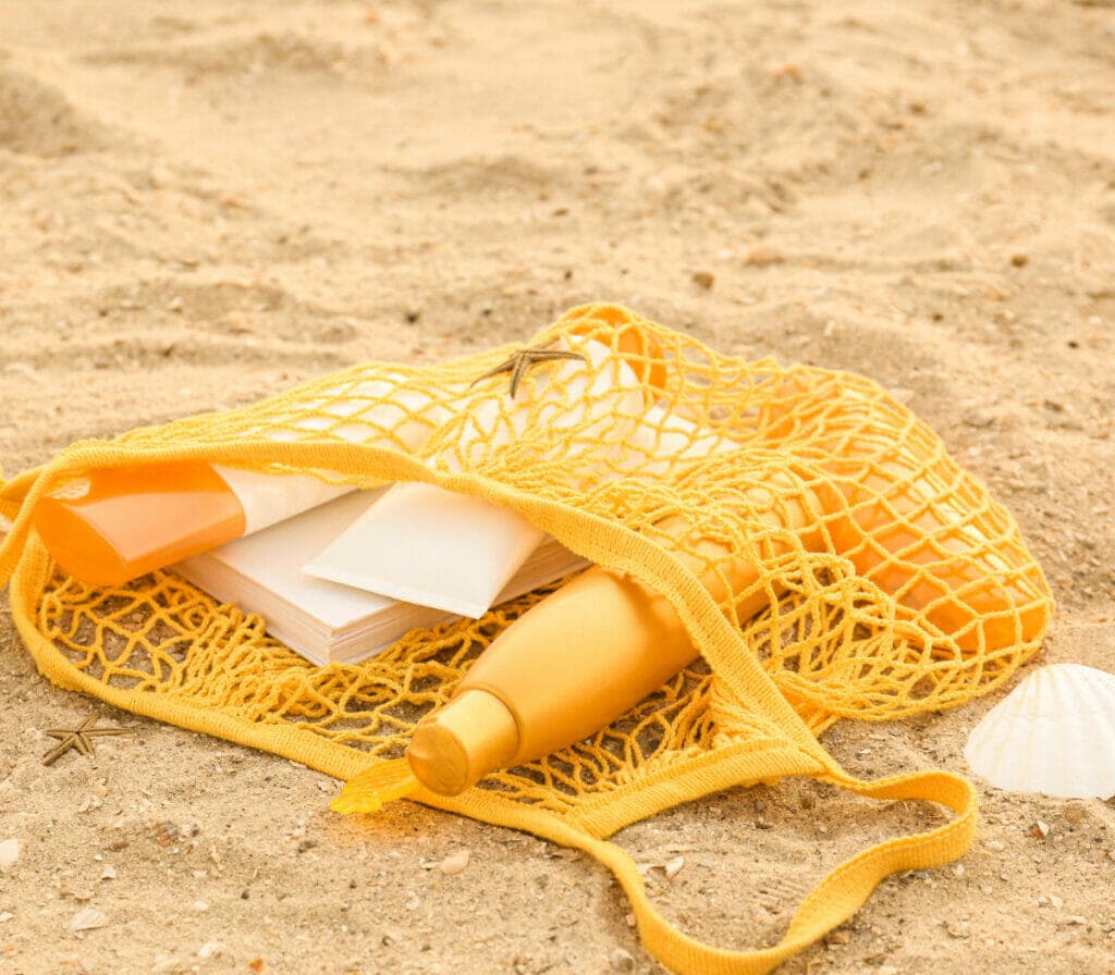 Yellow knit beach bag laying on sand holding sunscreen bottles 
