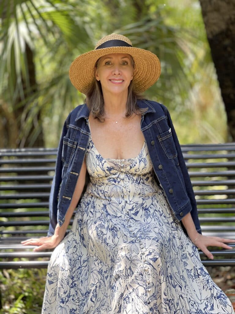 Nina Anders from Sharing A Journey Blog wears straw hat, denim jacket and blue and white sun dress.  Stylish Women Over 50