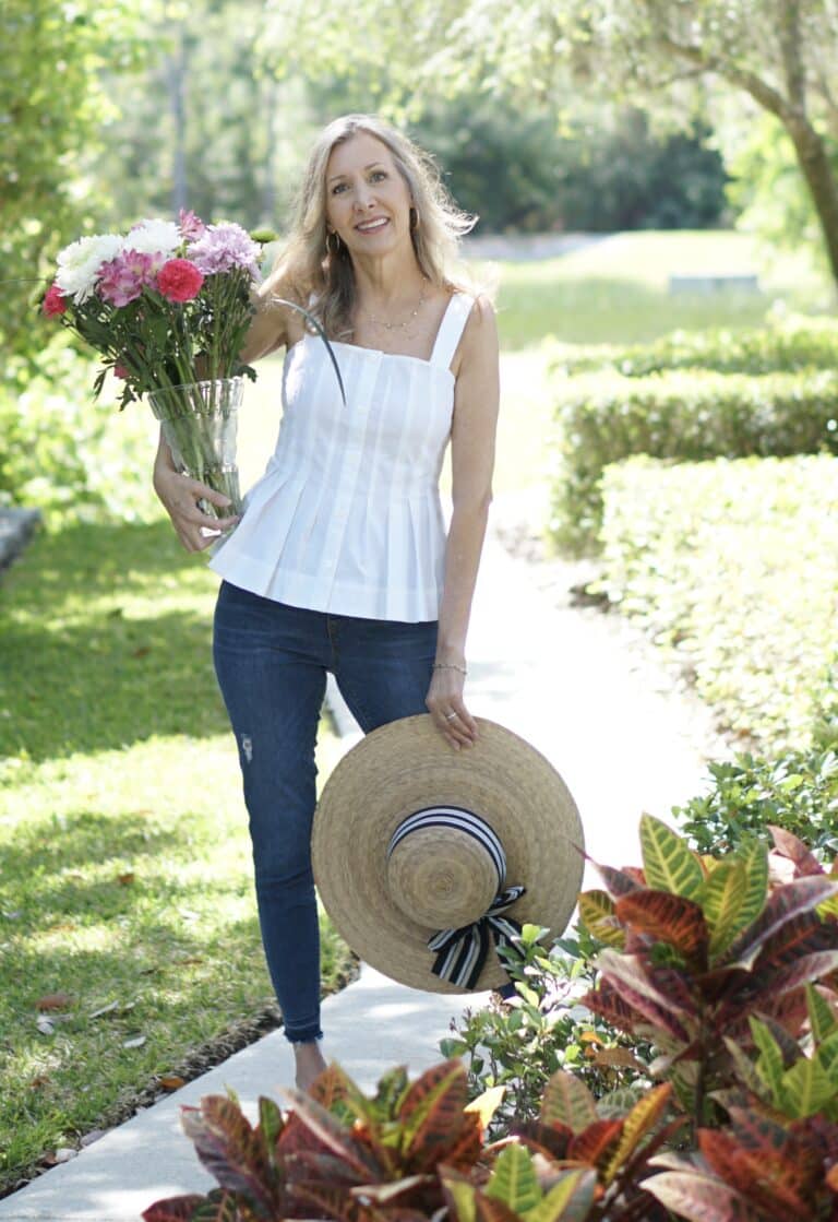 Nina Anders wearing white pin tuck blouse denim jeans holding flowers and a straw hat